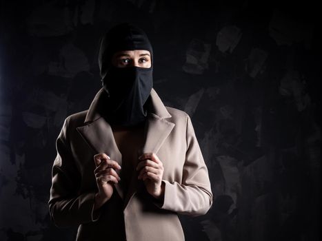 Portrait of a girl in a black balaclava and beige coat. Shot in the studio on a dark background.