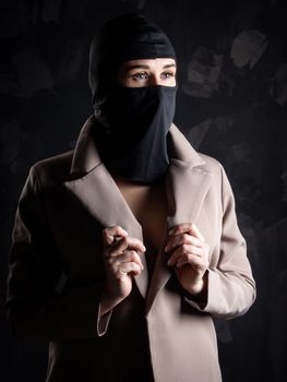 Portrait of a girl in a black balaclava and beige coat. Shot in the studio on a dark background.