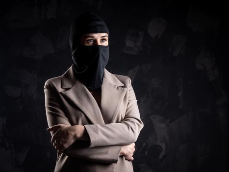 Portrait of a girl in a black balaclava and beige coat. Shot in the studio on a dark background.