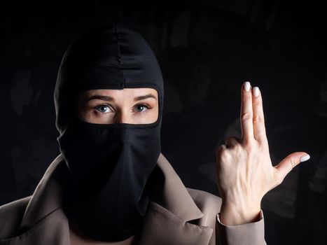 Portrait of a girl in a black balaclava and beige coat. Shot in the studio on a dark background.