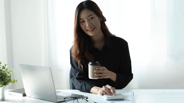 Smiling Asian businesswoman holding a coffee mug and laptop at the office. Looking at the camera..