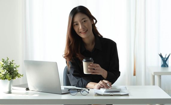 Smiling Asian businesswoman holding a coffee mug and laptop at the office. Looking at the camera..