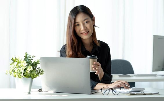 Smiling Asian businesswoman holding a coffee mug and laptop at the office. Looking at the camera..