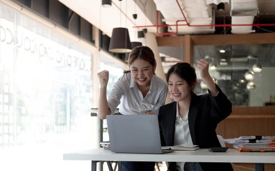 Two young Asian businesswomen show joyful expression of success at work smiling happily with a laptop computer in a modern office..