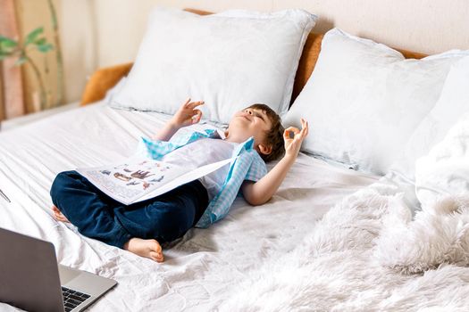 Distance learning online education. Caucasian happy boy lying with book studying home with laptop notebook and doing homework. Lying on bed with training books. Selective focus and blur background.