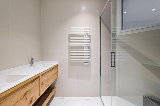 Modern bathroom with wooden base for white sinks. Radiator for warm water procedures in winter. Shower zone with glass door. Bathroom decorated with light tiles