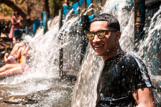 Latino teenager in sunglasses smiling bathing in a river with the water from a dam falling on his back