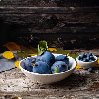 autumn background with ripe violet blue plums in white bowl on othe ld grey wooden table, side view