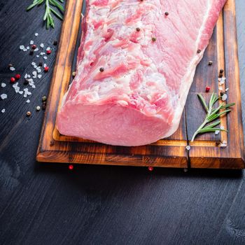 Big whole piece of pork meat with seasoning on chopping board on dark background with rosemary, seasonings, side view.