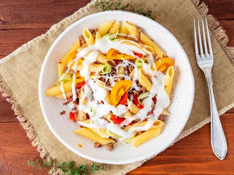 Penne pasta with yellow tomatoes, vegetables, mincemeat, white sauce on dark wooden background, top view, close up.