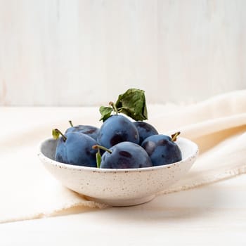 violet blue ripe plums in the white bowl on white wooden table with linen textile ranner tablecloth, side view