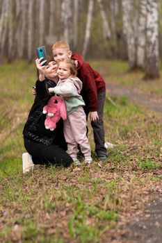 A mother with her son and daughter in a birch grove take a selfie near the path. they are smiling sweetly, and a pink monster bag is hanging around the girl's neck