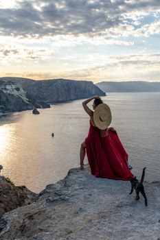 A girl with loose hair in a red dress and hat stands on the rocks above the sea. In the background, the sea and the rocks. The concept of travel