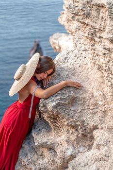 A girl with loose hair in a red dress and hat stands on the rocks above the sea. In the background, the sea and the rocks. The concept of travel