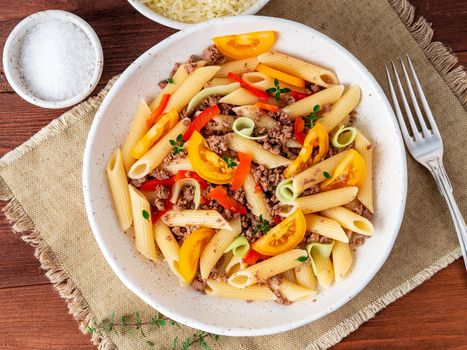 Penne pasta with yellow tomatoes, red and green vegetables, mincemeat on dark wooden background, top view, close up.