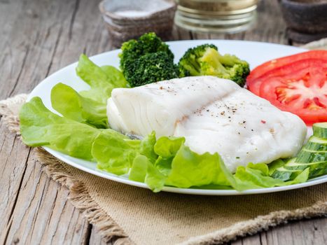 Steamed cod fish. Paleo, keto, fodmap healthy diet with vegetables on white plate on wooden table, side view.
