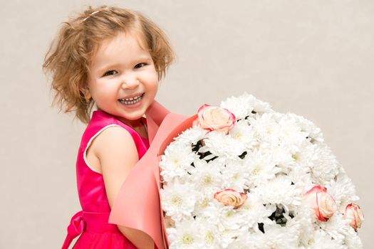 A little girl in a bright pink dress with curly hair is holding a huge bouquet of chrysanthemums and roses for her mother and smiling broadly and fervently. the baby will soon give it to her beloved mother