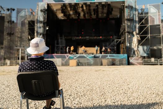in front of a huge open stage, where an open-air arts festival will be held on a huge empty platform in front of the stage, the director of the show sits on a chair and watches the rehearsal taking place on stage