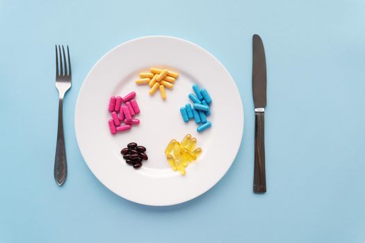 Colorful pills in a large white bowl along with a fork and knife on a blue background. Health concept