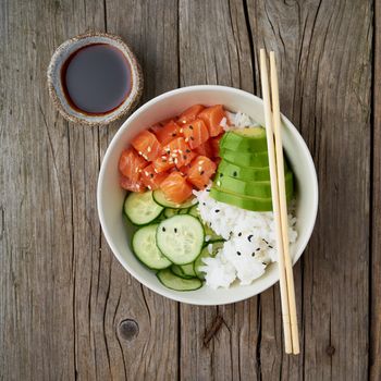 Salmon poke bowl with fresh fish, rice, cucumber, avocado with black and white sesame. Old wooden table. Food concept.
