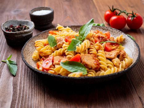 fusilli pasta with tomato sauce, chicken fillet with basil leaves on dark brown wooden background, cope space, side view.