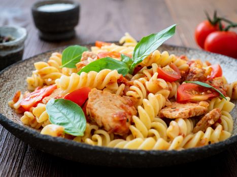 fusilli pasta with tomato sauce, chicken fillet with basil leaves on dark brown wooden background, cope space, side view.