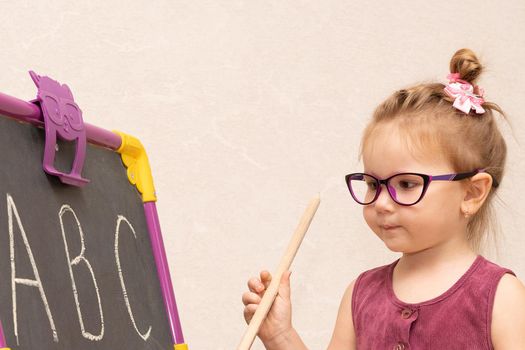 cheerful emotional girl of three years with a strict hairstyle like a teacher in a Burgundy dress and glasses. near the blackboard shows students. the letters in the alphabet....