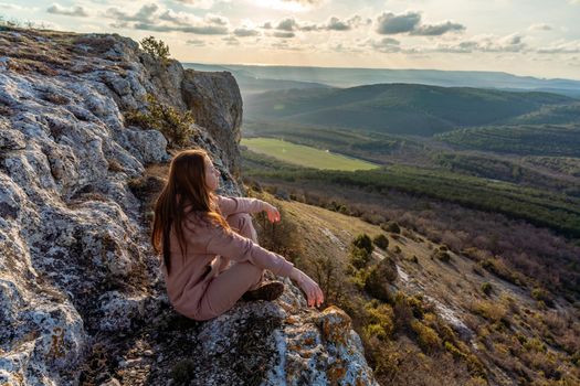 A girl sits on the edge of the cliff and looking at the sun valley.Woman enjoying a sunset or sunrise from the top of the mountain in summer.The traveler girl see the sunrise at the peak of mountain