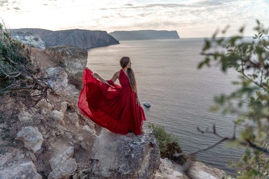 A girl with loose hair in a red dress stands on a rock rock above the sea. In the background, the sea and the rocks. The concept of travel.