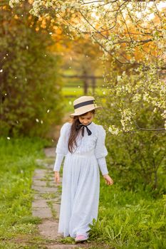 A beautiful girl in a white dress and a hat walks under flowering trees in the spring