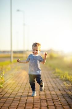 Cheerful little boy with striped shirt runs down the sidewalk. Sunny summer evening, backlighting.