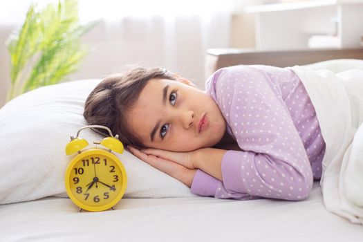 A cute disgruntled brunette in purple polka dot pajamas lies on a white pillow, on the bed, in the morning light, there is a yellow alarm clock nearby. copy space