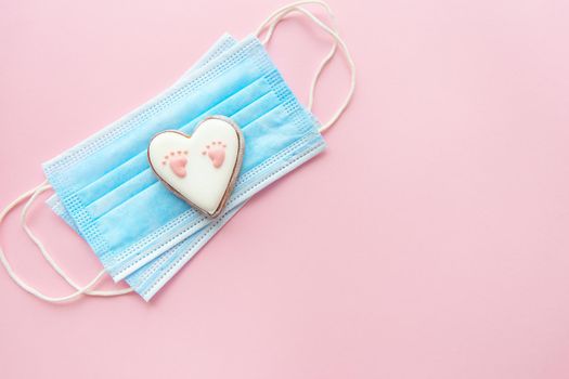 Disposable medical mask and heart with baby footprint on pink background. Childbirth during the coronavirus pandemic