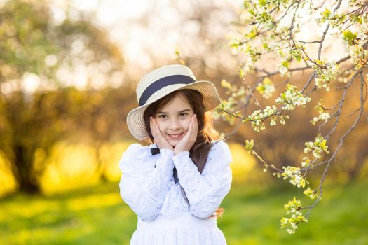Portrait of adorable little girl in a white dress and hat, stands in the park, under flowering trees, in spring