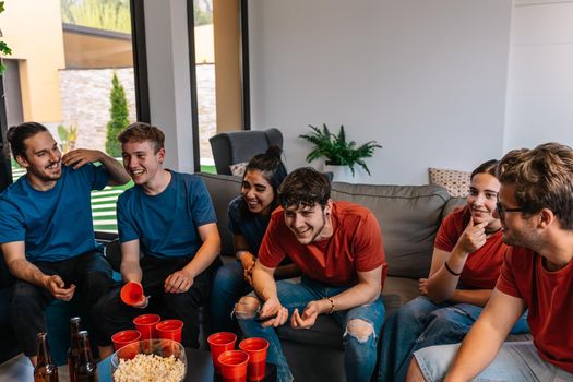 friends watching a football match on television and competing for their team. group of young people at a party. living room sofa, inside the house, natural light, trumpets to cheer for their team, food and drinks.