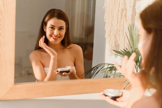 A young woman applies moisturizer to her face in her sun bath. Daily routine, hydration and skin care concept.