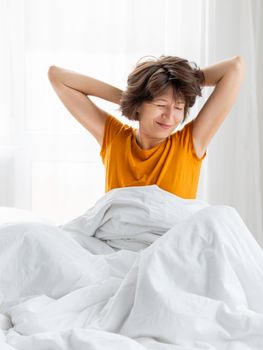 Sleepy woman in yellow pajama is stretching in bed, covered with white blanket. Waking up early in morning. Woman gets enough sleep.
