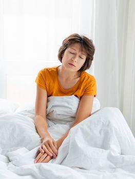 Sleepy woman is sitting in bed, completely covered with white blanket. It's hard to wake up early in morning. Woman does not get enough sleep.