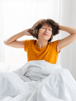 Sleepy woman in yellow pajama is stretching in bed, covered with white blanket. Waking up early in morning. Woman gets enough sleep.