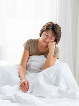 Sleepy woman is sitting in bed, completely covered with white blanket. It's hard to wake up early in morning. Woman does not get enough sleep.