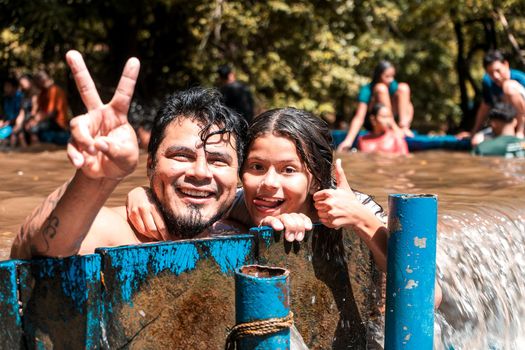 Father and daughter looking at the camera and making a peace gesture with their hands and fingers submerged in a dam in a lake in Masachapa, Nicaragua.