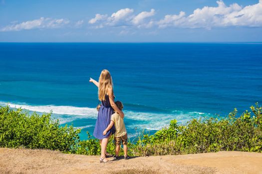 Mom and son travelers on a cliff above the beach. Empty paradise beach, blue sea waves in Bali island, Indonesia. Suluban and Nyang Nyang place. Traveling with kids concept.