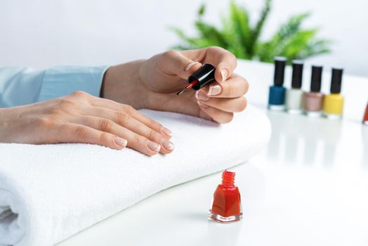 Woman giving herself elegant manicure at home. Closeup of beautiful female hand applying red nail polish. Colorful nail polish bottles on table. Nail care and beautician procedure. Stylish nail art