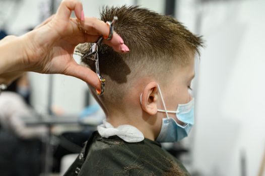 A teenage boy gets a haircut in a barbershop during a pandemic, a haircut in the salon, a client and a hairdresser in masks, a baby haircut with scissors.
