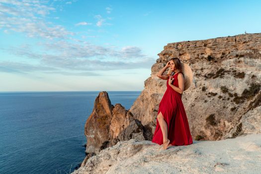 A girl with loose hair in a red dress stands on a rock rock above the sea. In the background, the sea and the rocks. The concept of travel.