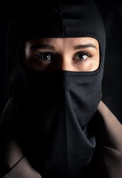 Portrait of a girl in a black balaclava and beige coat. Shot in the studio on a dark background.