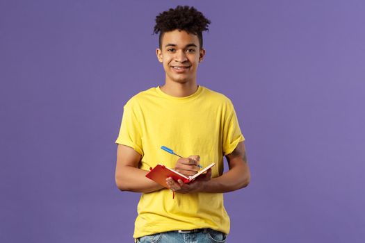 Portrait of young hispanic male student studying online courses, writing down lecture, making personal schedule or taking notes in planner, look camera enthusiastic and interested, purple background.