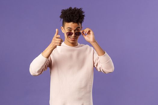 Portrait of cheeky handsome young man wearing sunglasses, take them off and show thumb-up, like or approval, recommendation sign, smirk satisfied, saying all good, alright, purple background.