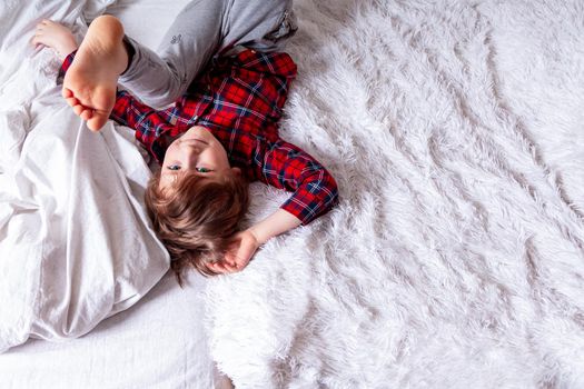 Happy little curly kid boy with smiley face lying on the white bed at home and legs up. Stay at home quarantine coronavirus COVID-19 pandemic prevention. With space for text.