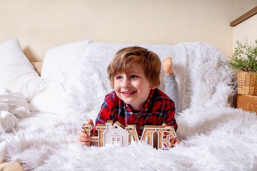 Happy little curly kid boy with smiley face lying on the bed at home. Stay at home quarantine coronavirus COVID-19 pandemic prevention. Wooden house inscription home.